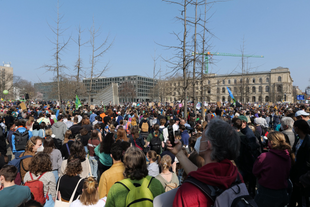 Eine große Menschenmenge mit Schildern und Taschen vor einem Gebäude mit Bäumen und einem klaren Himmel, was auf eine Klimaprotest demonstration hinweist.