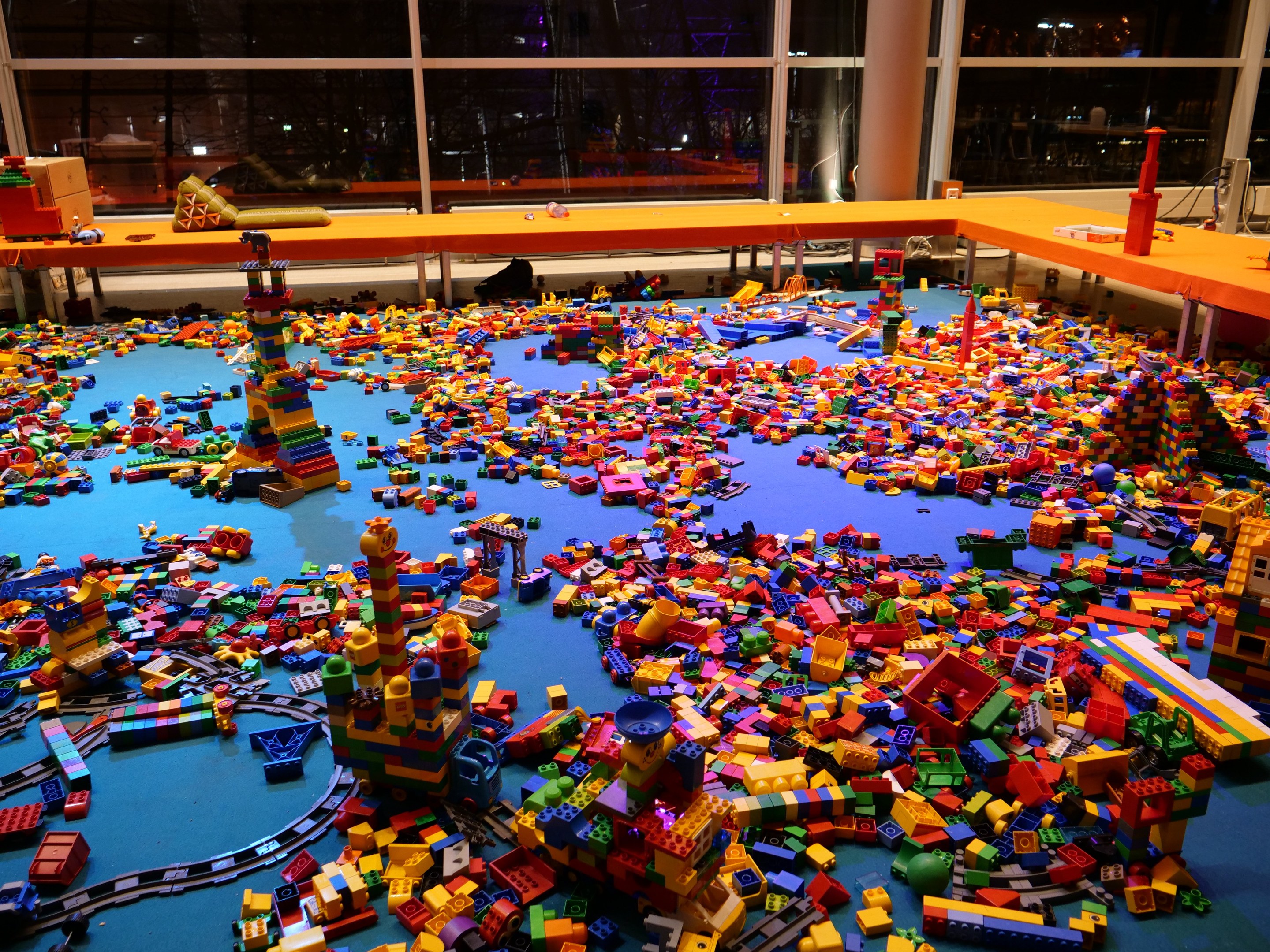 Ein großer Raum mit farbigen Lego-Bauklötzen, die auf dem Boden verstreut sind, umgeben von Tischen und Säulen, mit Glasfenstern, die Menschen und Lichter im Hintergrund zeigen.