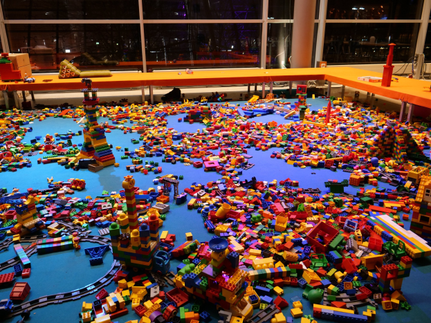 Ein großer Raum mit farbigen Lego-Bauklötzen, die auf dem Boden verstreut sind, umgeben von Tischen und Säulen, mit Glasfenstern, die Menschen und Lichter im Hintergrund zeigen.