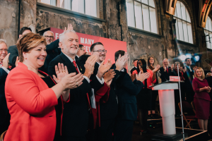 Eine Gruppe von Menschen, die vor einem Publikum klatschen, mit einem Podium, einem Mikrofon und einer Tafel mit Text auf der rechten Seite und Stühlen, einem Banner, einer Wand, Fenstern und Lichtern im Hintergrund.
