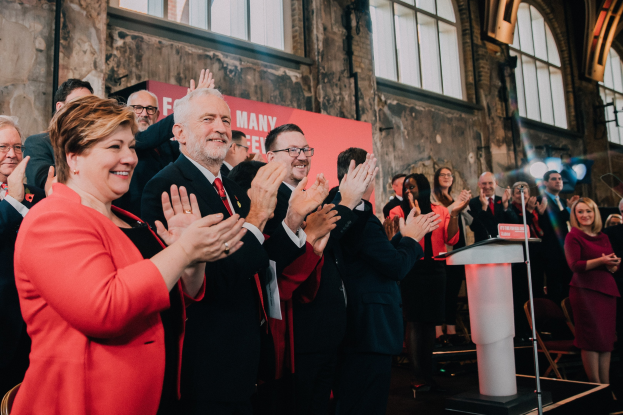 Eine Gruppe von Menschen, die vor einem Publikum klatschen, mit einem Podium, einem Mikrofon und einer Tafel mit Text auf der rechten Seite und Stühlen, einem Banner, einer Wand, Fenstern und Lichtern im Hintergrund.