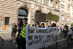 Eine Gruppe von Menschen marschiert auf einer Straße in Berlin, hält Plakate und Banner, während einige Fahrráder fahren, vor einem Gebäude mit Überdachungen, Pfeilern, Skulpturen und Bäumen.