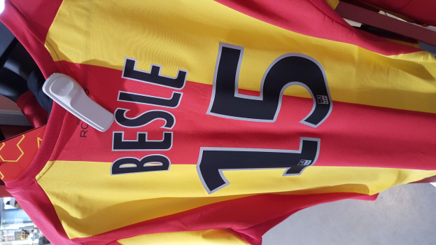 Rotes und gelbes Fußballtrikot mit bedrucktem Text und Nummern, das an einer Ladengestell hängend mit anderen Gegenständen und dem Hintergrund im Hintergrund zu sehen ist.