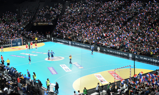 Eine Gruppe von Menschen, die Handball in einem Stadion mit einem Netz auf dem Platz und Zuschauern in den Rängen spielen, während Fotografen das Spiel aufnehmen.