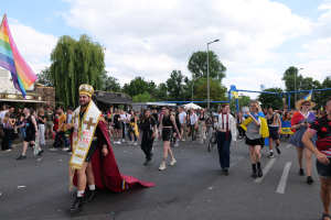 Eine Gruppe von Menschen marschiert während der Gay Pride Parade 2018 die Straße entlang, einige tragen eine Regenbogenflagge und Musikinstrumente, mit Laternenmasten, Bäumen, Schuppen und einem bewölkten Himmel im Hintergrund.