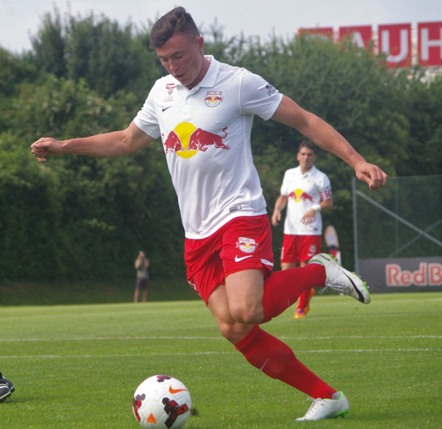 Ein professioneller Fußballspieler, der einen Fußball auf einem Rasenfeld mit "RB Leipzig" im Hintergrund unter einem klaren blauen Himmel kickt.