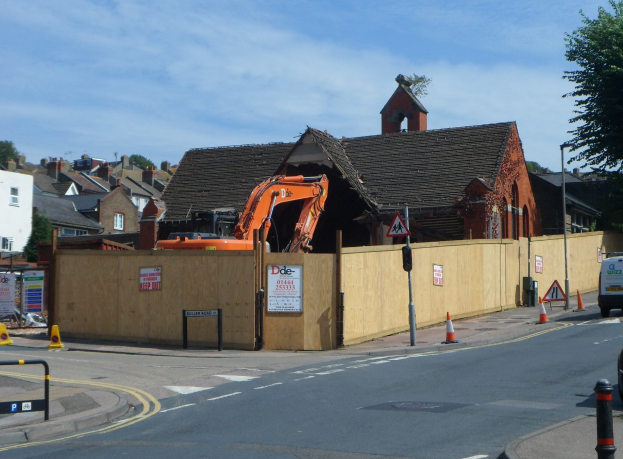 Straße mit Fahrzeugen, Verkehrskegeln, Pfählen, Schildern und einem Zaun; Bagger und abgerissenes Gebäude am Straßenrand; Gebäude, Bäume, Wolken und Himmel im Hintergrund.
