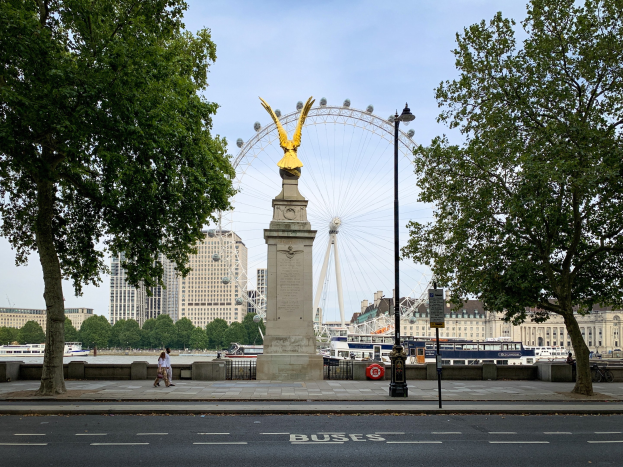 Das London Eye mit einer Statue auf einem Podest im Vordergrund, eine Straße, Bäume, ein Laternenpfahl, Menschen auf einem Fußweg, Schiffe auf dem Wasser, Gebäude und der Himmel im Hintergrund.