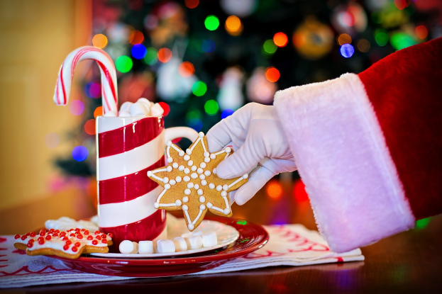 Eine Person mit einem Santa Claus Hut hält einen Becher heißer Schokolade und einen Bonbonstab in der Hand und hat einen Teller mit Plätzchen und ein Tuch auf dem Tisch vor sich. Im Hintergrund sieht man einen Weihnachtsbaum und eine Wand.