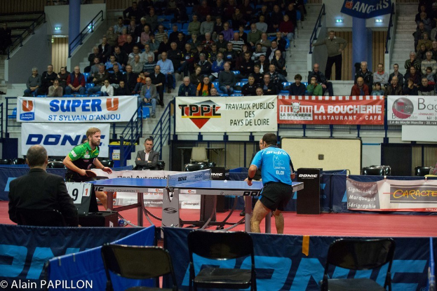 Indoor-Tischtennis-Match mit zwei Spielern, die gegeneinander antreten, einigen stehenden Zuschauern, sitzenden Publikum, leeren Stühlen und Werbetafeln in einer Stadionatmosphäre.