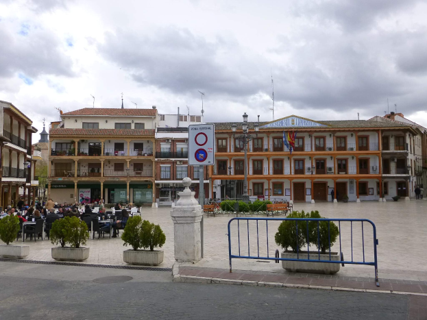 Ein belebter Stadtplatz mit Menschen auf Stühlen sitzend und stehend, umgeben von Topfpflanzen, Metallabsperrungen, Straßenmasten mit Fahnen, ein Schild, Gebäude mit Fenstern und einem bewölkten Himmel.