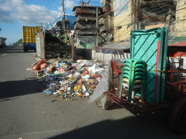 Ein Lastwagen neben einem Haufen Müll auf einer Straße geparkt, mit einem Wagen mit Plastikstühlen rechts daneben und Gebäuden, Strommasten, Bäumen und einem bewölkten Himmel im Hintergrund.