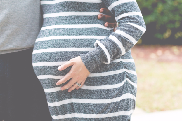 Schwangere Paare stehen zusammen mit den Händen auf dem Bauch, tragen Schwangerschaftskleidung, umgeben von Bäumen.