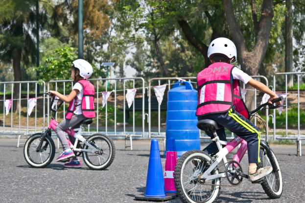 Zwei junge M├Ądchen in rosa und wei├čen Kürzen und wei├čen Helmen fahren auf Fahrr├Ądern eine Stra├če mit Verkehrskegeln hinunter, mit einer blauen Trommel, einem Gel├Ąnder, B├Ąumen, einem Pfahl und Himmel im Hintergrund.