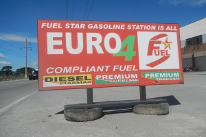 Eine Tankstelle mit einem "Euro4 Fuel"-Schild im Vordergrund, Fahrzeuge auf der Straße, Strommasten mit Käble, Bäume, ein Gebäude auf der rechten Seite und sichtbarer Himmel.