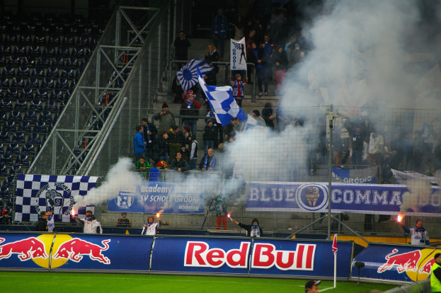 Eine Gruppe von Menschen auf einem Fußballfeld, die Fahnen und Banner schwenken, mit Rauch auf dem Boden, umgeben von einem Metallzaun und leeren Stadionstühlen.