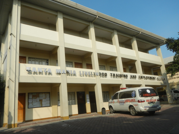 Santa Maria Lebensmittelpunkt- und Arbeitsplatz-Trainingszentrum mit Fenstern, Türen und Text, umgeben von Bäumen und Fahrzeugen auf der Straße unter einem klaren blauen Himmel.