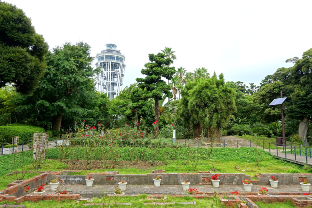 Ein Park mit einem Turm im Hintergrund, umgeben von grünem Gras, Pflanzen, Bäumen, Blumentöpfen, einer Straße mit Geländern und einem Pfahl mit einer Tafel, unter einem sichtbaren Himmel.