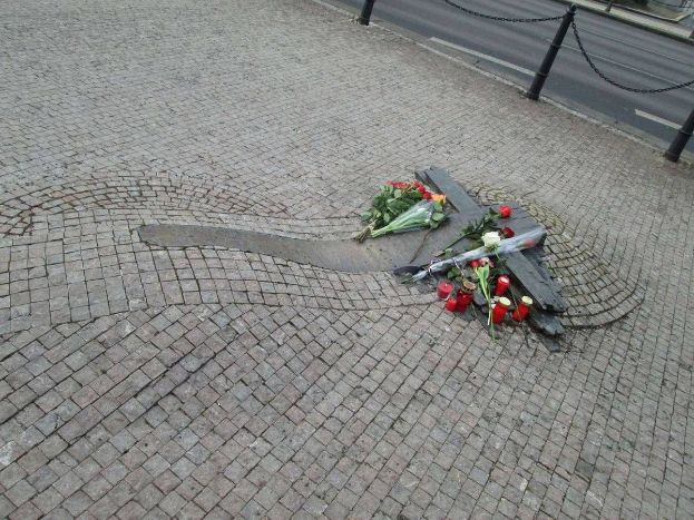 Gedenkstätte mit Blumen, Kerzen und verstreuten Buketts am Straßenrand, umgeben von Bäumen und Gebäuden, mit Fahrzeugen und angeketteten Pfählen im Hintergrund.
