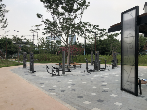 Outdoor-Park mit verschiedenen Fitnessgeräten, Bänken, Bäumen, Pflanzen, Gras, Pfosten, Lichtern, Drähten und Gebäuden vor einem Himmel-Hintergrund.
