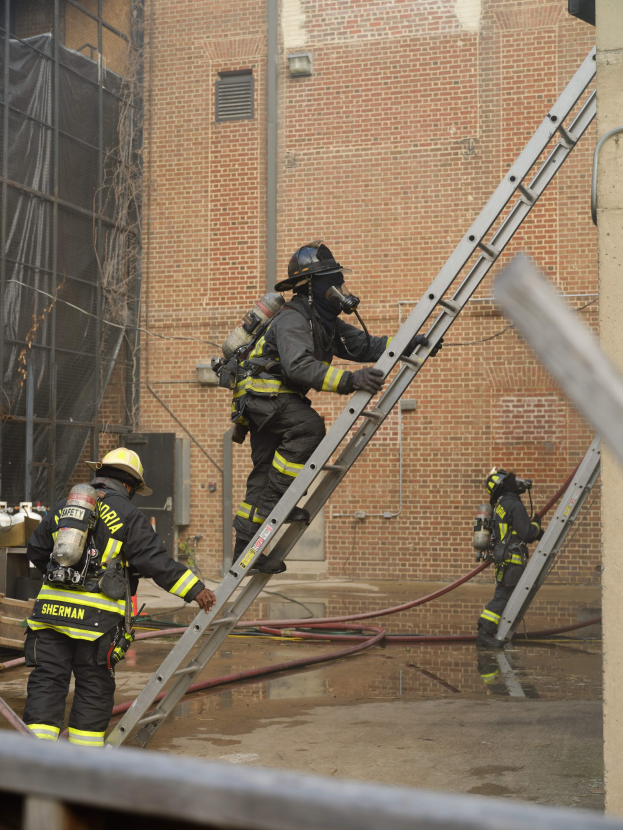 Feuerwehrleute mit Helmen und Luftzylindern klettern an einer Leiter vor einem Backsteingebäude mit Rohren auf dem Boden und einer Metallstange am Boden, mit einem weiteren Gebäude mit Fenstern und Netz im Hintergrund.