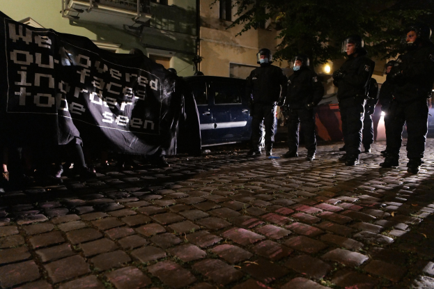 Eine Gruppe von Polizisten in Helmen steht auf einer Kopfsteinpflasterstraße bei Nacht, mit einem Banner, mit Fahrzeugen, Gebäuden, Bäumen und Straßenbeleuchtung im Hintergrund.