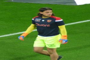 Ein Mann in einem blauen T-Shirt und gelben Shorts läuft auf einem Fußballfeld, die Arme ausgestreckt und mit entschlossener Miene, mit einem Tuch oben auf dem Bild.