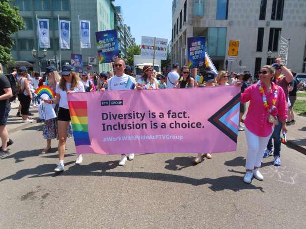 Eine Gruppe von Menschen marschiert auf einer Straße, hält ein Banner mit der Aufschrift "Diversität ist eine Tatsache, Inklusion ist eine Wahl" in den Händen, einige tragen Mützen und Schutzbrillen, andere zusätzliche Banner, vor dem Hintergrund von Gebäuden, Bäumen, Laternenmasten und einem klaren blauen Himmel.