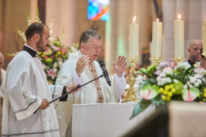 Bischof der Kirche des Heiligen Grabes in Priestergewand vor einem Rednerpult mit Mikrofon, umgeben von Menschen, mit einem Tisch mit Kerzen und Blumen rechts, Blumensträußen und einem Bleiglasfenster im Hintergrund.