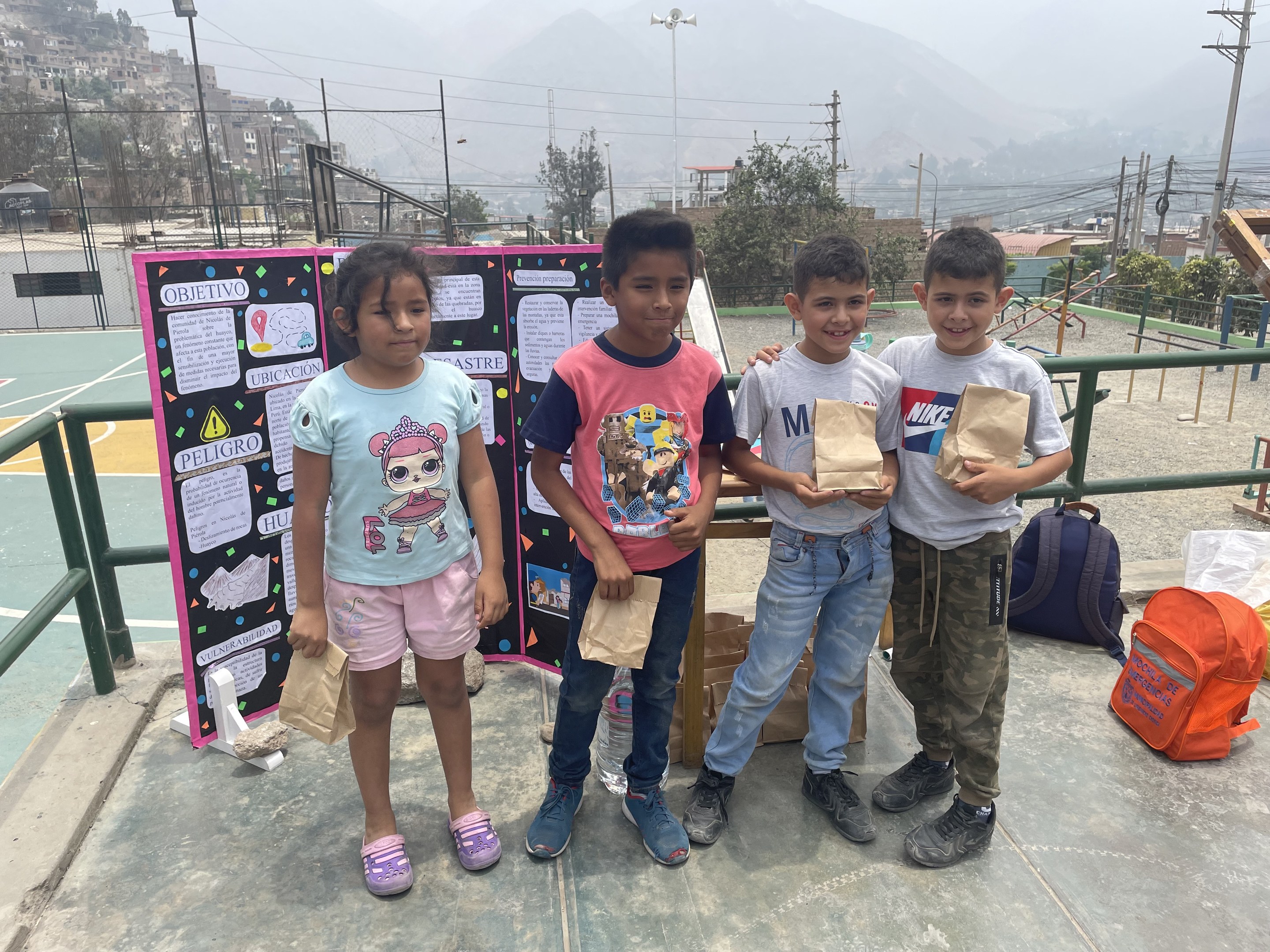 Gruppe von Kindern mit Papiertüten auf einem Basketballfeld mit Materialien auf dem Boden, eine Tafel mit Text links und Zaun, Pfosten und Gebäude im Hintergrund.