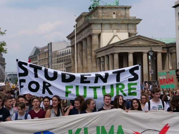 Gruppe von Schülern marschiert in Berlin mit einem bunt bemalten "Schüler für die Zukunft"-Schild vor Gebäuden, Bäumen und Himmel.