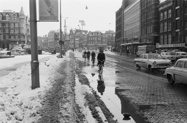 Schwarzes und weißes Foto von Radfahrern und Autos auf einer verschneiten Straße, die von Gebäuden gesäumt ist, verschneiter Boden, ein Schild im Vordergrund und der Himmel im Hintergrund.