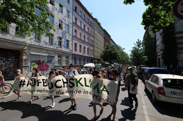 Menschen mit einem Transparent auf der Straße, mit parkenden Autos auf der rechten Seite, einem Radfahrer auf der linken Seite und Gebäuden, Bäumen und einem Schild im Hintergrund während einer Demonstration in Berlin.