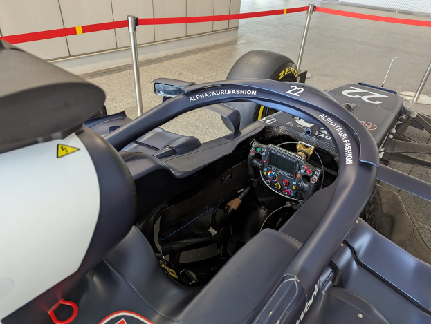 Cockpit eines Formel-E-Autos in einer Ausstellung mit einem Lenkrad im Vordergrund, einer Wand im Hintergrund und dekorativen Bändern um die das Auto herumstehenden Pfosten.