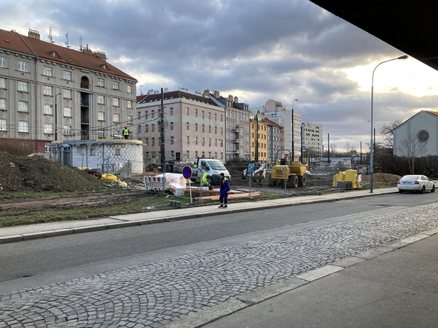 Eine Straße in einer Stadt mit parkenden Autos, Gebäuden, Laternen, Bäumen und einem bewölktem Himmel, mit einer Straßensperre mit Fahrzeugen und Fußgängern im Vordergrund.