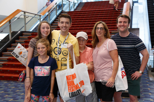 Gruppe von Menschen, die vor einer Rolltreppe stehen und Taschen halten, während sie lächeln. Im Hintergrund sind Treppen, Geländer und eine Wand zu sehen.