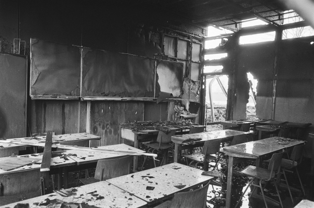 Schwarz-weiß-Foto eines verlassenen Klassenzimmers mit verbrannten Wänden, zerbrochenen Fenstern und verstreuten Tischen und Stühlen.