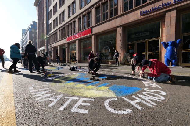 Menschen sitzen vor einem Gebäude mit Fenstern und Namensschildern während einer Klimaprotest in Berlin, umgeben von Flaschen und anderen Gegenständen, mit Bäumen und einem klaren blauen Himmel im Hintergrund.
