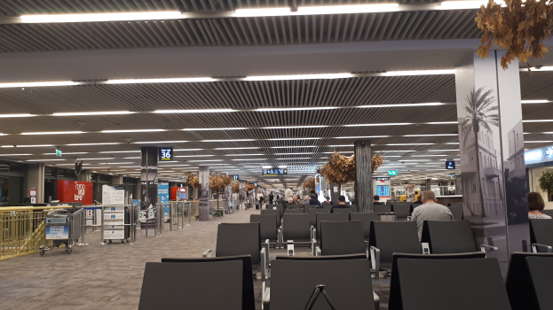 Ein leerer Flughafen mit Reihen von Stühlen, einige besetzt von Menschen, Säulen, Texttafeln, Deckenleuchten, Geländern und verstreuten Gepäckwagen.