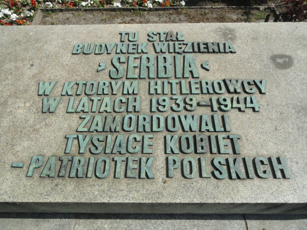 Gedenkstein mit einer Tafel vor einem Gebäude umgeben von blühenden Pflanzen.