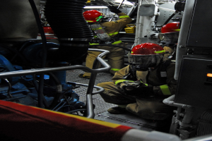 Feuerwehrleute in Helmen und Schutzbekleidung bei der Arbeit an einem Feuerlöschwagen mit Rohren und Gegenständen im Hintergrund, unter einem Licht an der Spitze.