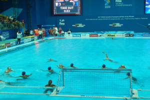 Eine Gruppe von Menschen, die Wasserball in einem Pool mit einem Netz spielen, umgeben von Zuschauern, Tafeln mit Text und Logos, Trägern und einem Bildschirm mit Zahlen und Text bei den 2015 FINA-Weltmeisterschaften in Kazan.
