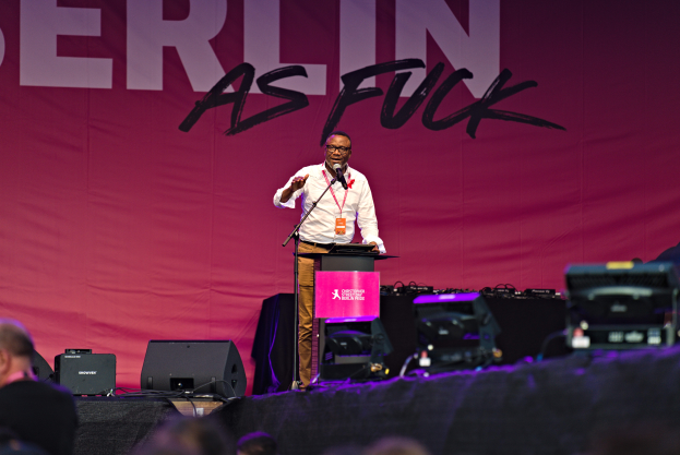 Ein Mann in einem weißen Hemd und braunen Hosen steht auf einer Bühne und hält ein Mikrofon in der Hand, mit einem pinken Banner hinter ihm, auf dem 'Berlin as Fuck' steht und Lautsprecher auf der Bühne.