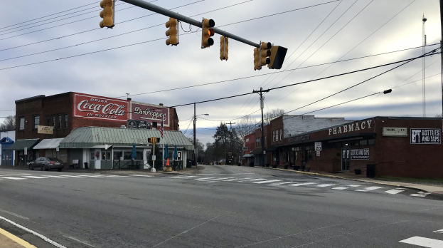 Eine städtische Straße mit einer Ampel an einer Kreuzung, Fahrzeuge auf der Straße, Gebäude mit Schildern, Strommasten mit Kabeln, Bäume und ein bewölkter Himmel im Hintergrund.