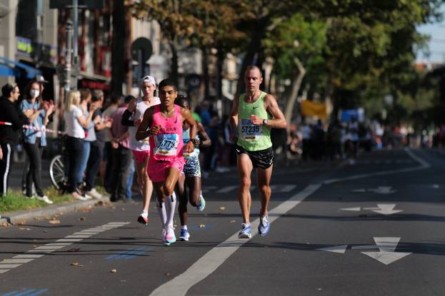 Gruppe von Läufern bei einem Marathon auf einer Stadtstraße mit Zuschauern auf der linken Seite, unscharfer städtischer Hintergrund mit Bäumen, Gebäuden und einem Fahrrad.
