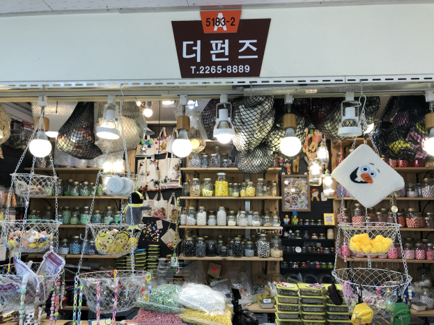 Ein unordentlicher Laden in Seoul zeigt verschiedene Gegenstände wie Körbe, Lampen, Flaschen und Spielzeuge auf Regalen, mit einer Tafel oben, die darauf hinweist, dass es ein top-bewerteter koreanischer Shop ist.