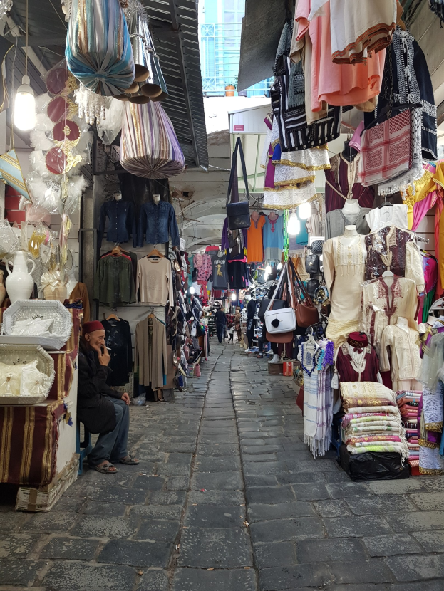Ein belebter Straßenmarkt in der alten Stadt Jerusalem mit Ständen, die Kleidung, Taschen, Lampen und andere Artikel verkaufen, umgeben von Menschen, die durch die Gassen schlendern und ein Gebäude im Hintergrund.