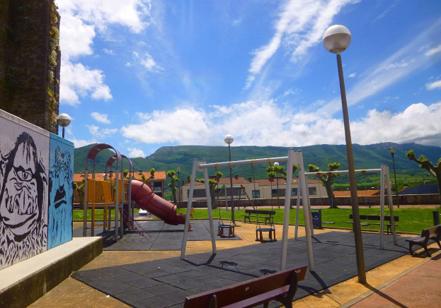 Ein Spielplatz mit Schaukeln, einer Rutsche, Bänken, Pfosten, Lichtern, einer bemalten Wand, Gras, Bäumen, Gebäuden, fernen Höhen und einem bewölktem Himmel.