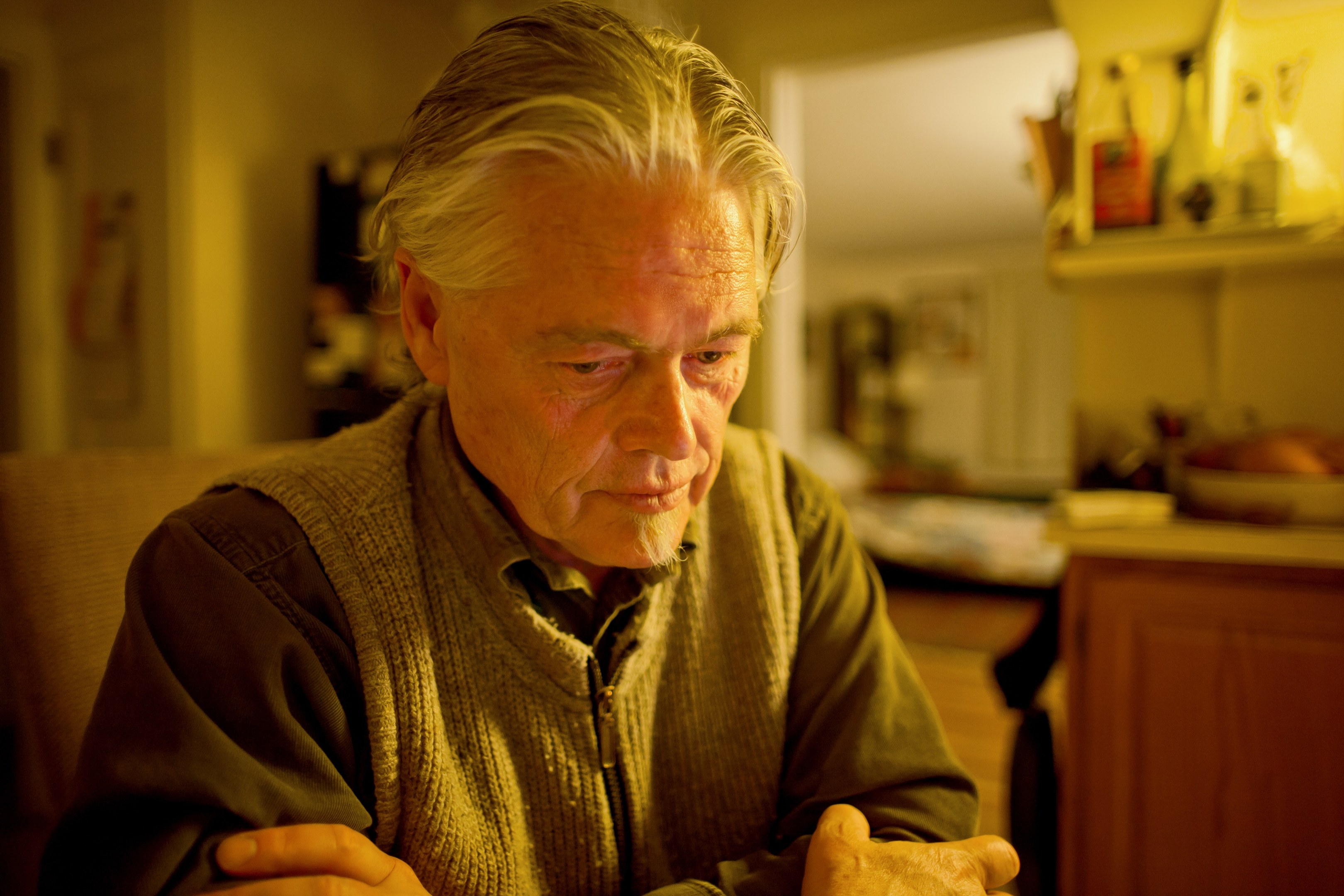 Ein älterer Mann sitzt an einem Tisch mit einem Teller Essen, mit einem Schrank voller Gegenstände rechts daneben und gerahmten Gegenständen und einer Tür im Hintergrund.