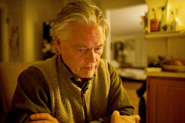 Ein älterer Mann sitzt an einem Tisch mit einem Teller Essen, mit einem Schrank voller Gegenstände rechts daneben und gerahmten Gegenständen und einer Tür im Hintergrund.